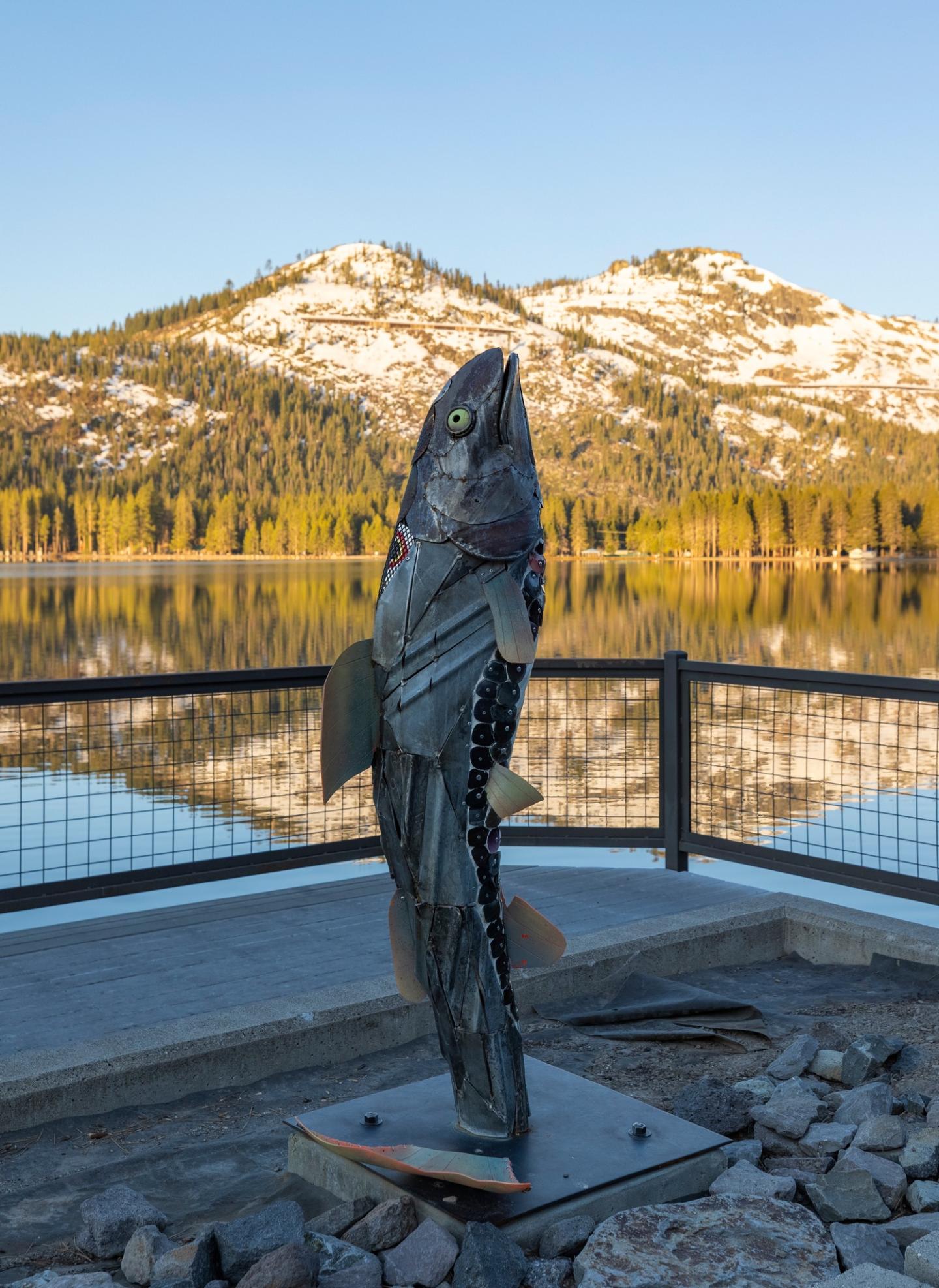 Metal fish sculpture by a lake with mountains in the background.