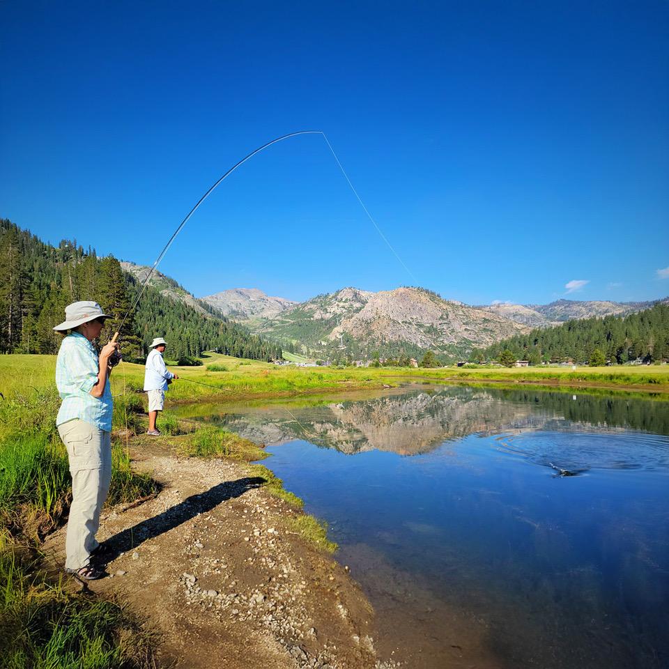 Two people fishing by a calm lake, surrounded by mountains and trees.