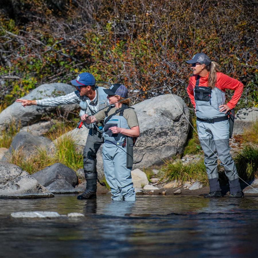 Three people in waders stand in a river, one pointing ahead, surrounded by rocks and bushes.