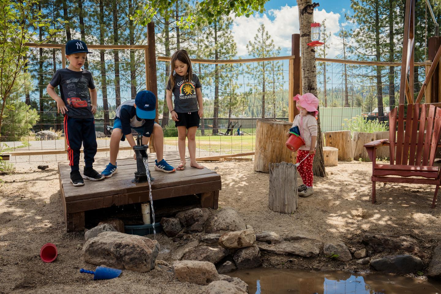 Children play by a pond with rocks and a sandbox in a forested outdoor setting.