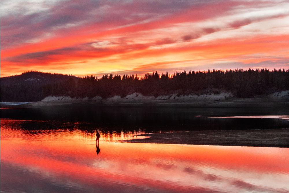 Fiery sunset over a calm lake with silhouetted trees and a lone figure.