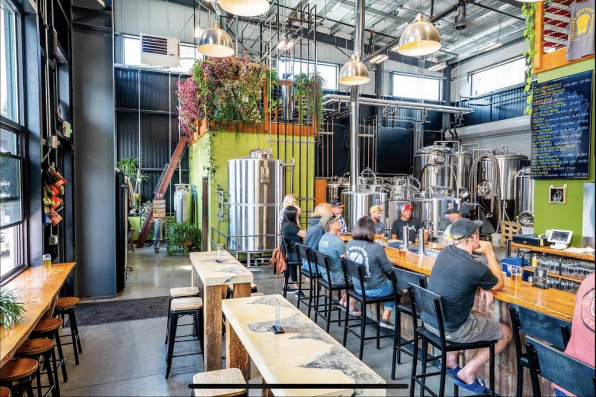 Modern brewery interior with people sitting at a bar, stainless steel tanks in the background.