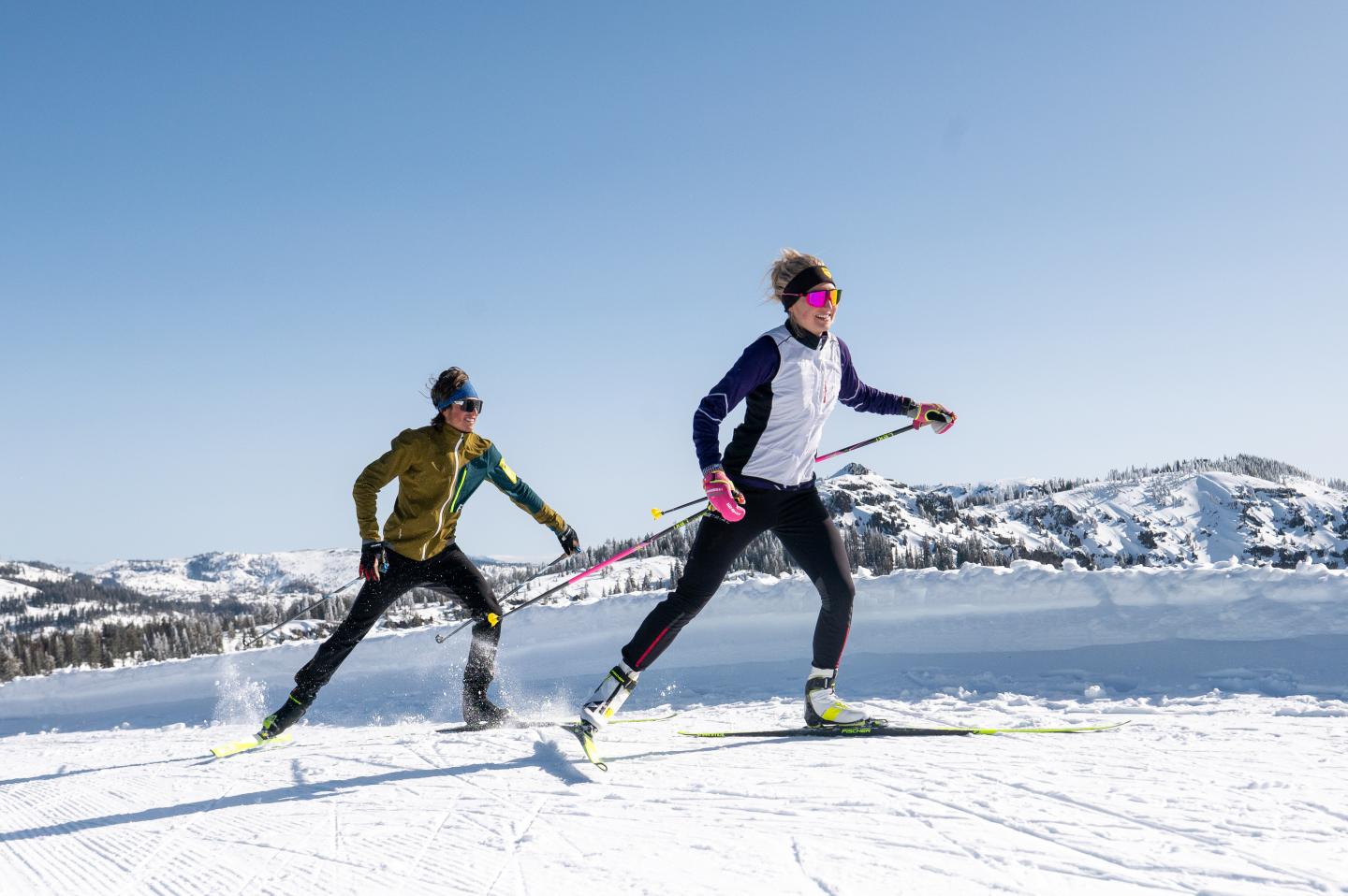 Two people cross-country skiing on a snowy landscape under a clear blue sky.