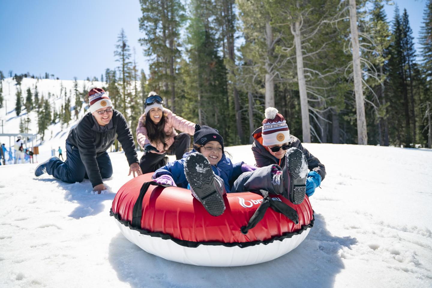 Two adults push two children on a snow tube under a clear sky.