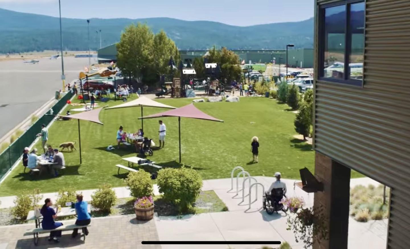 Outdoor gathering with people on a sunny lawn, shaded tables, and mountain view.
