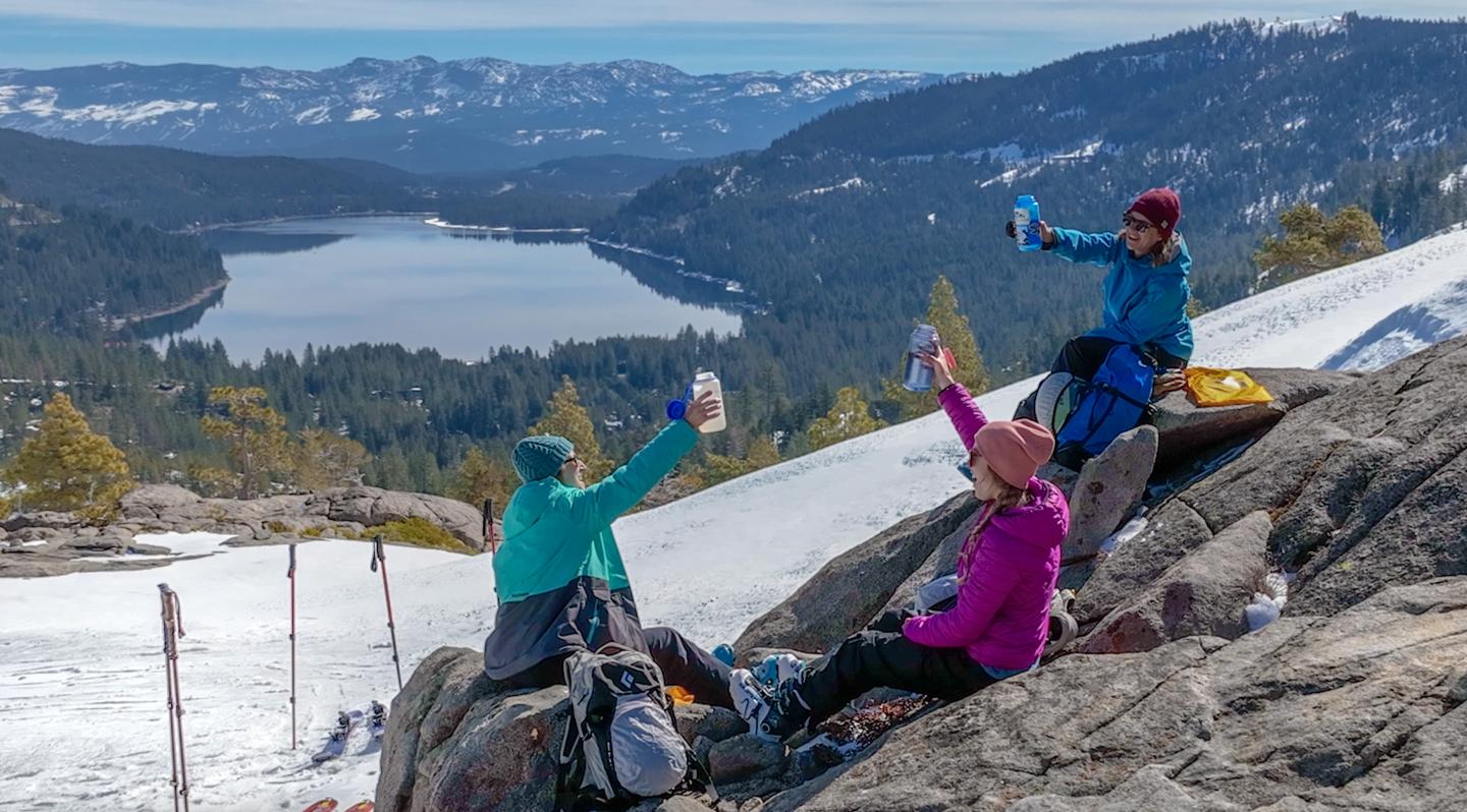 Three people sit on a snowy hillside, raising drinks near a scenic lake and mountains.