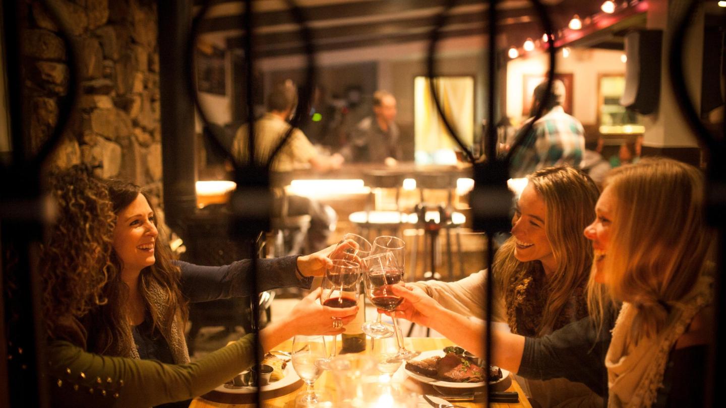 Three women toasting with wine glasses in a cozy, warmly lit restaurant.