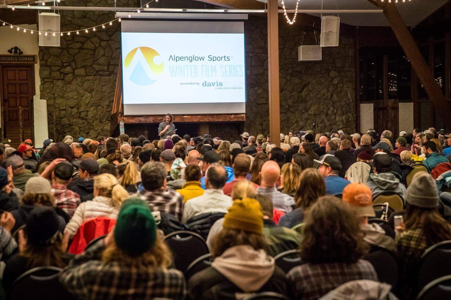Audience in a crowded room watches a presentation on a large screen.