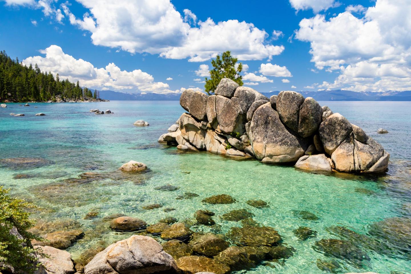 Lake Tahoe, Clear lake with rocks, surrounded by trees and blue sky with clouds.
