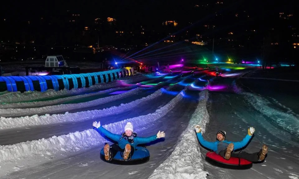 Snow tubing at night with colorful lights and two people descending a snowy slope.