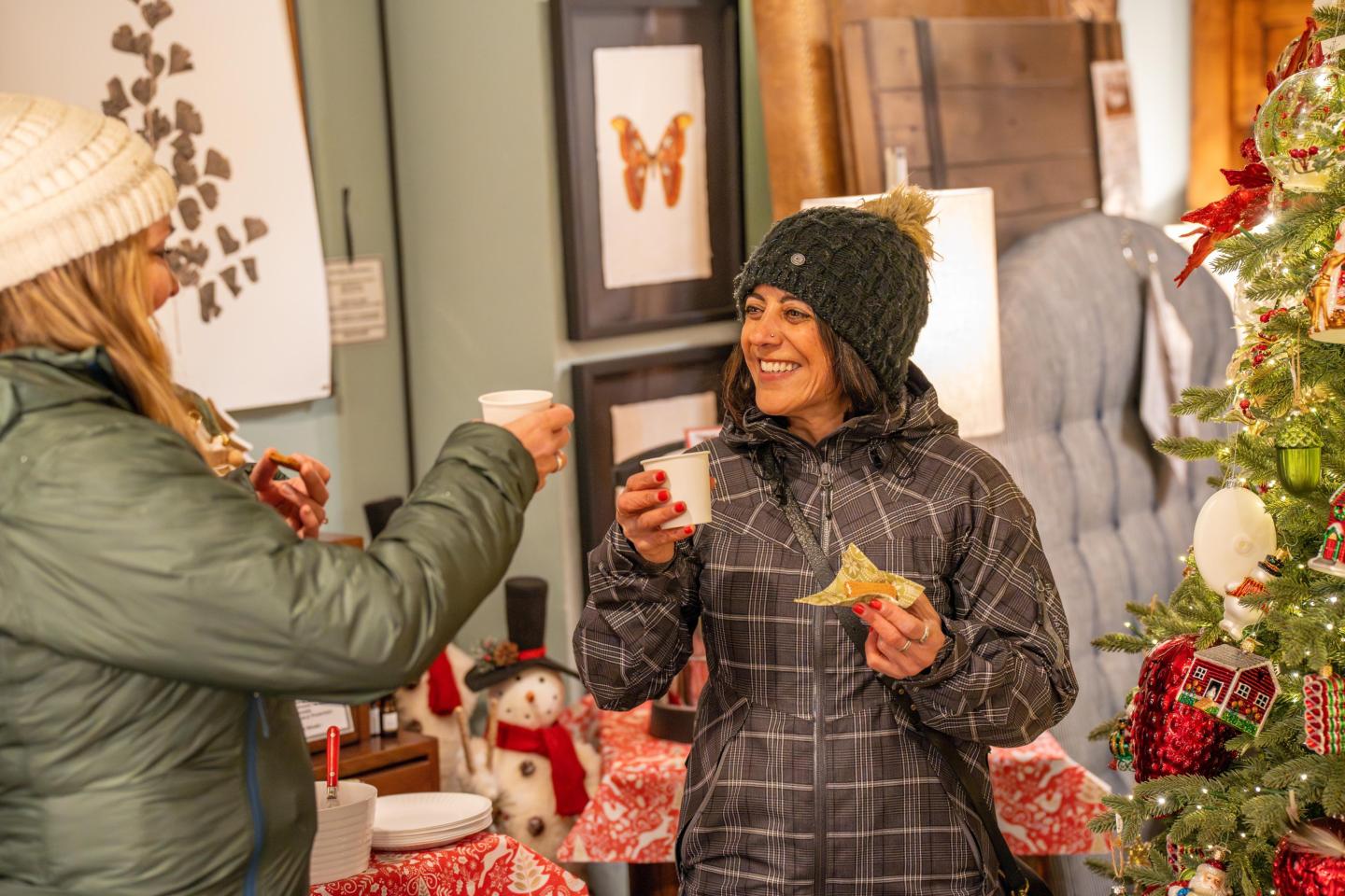 Two women in winter clothes enjoying drinks and snacks at a festive indoor setting.