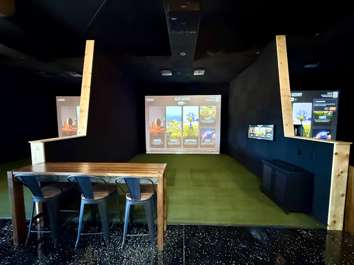 Indoor golf simulator room with screens and green flooring.