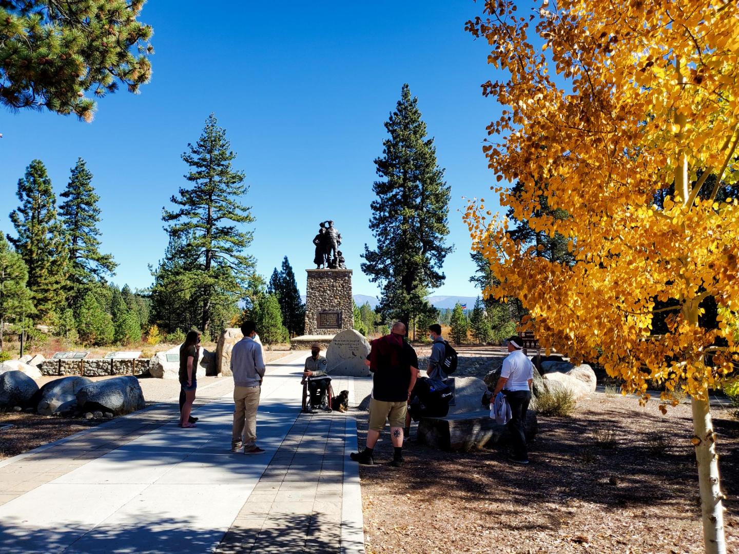 People gather near a monument with autumn trees and clear blue sky.