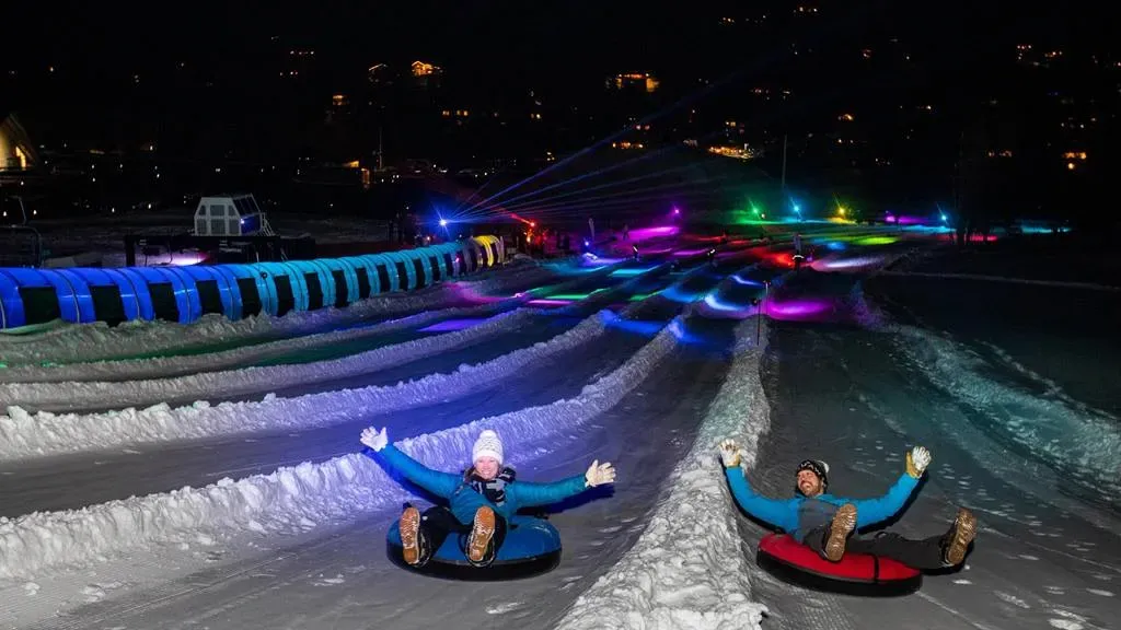 Snow tubing at night with colorful lights and two people descending side by side.