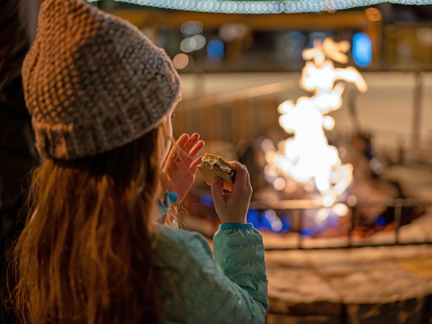 Girl in a beanie roasting marshmallows by a fire pit at night.