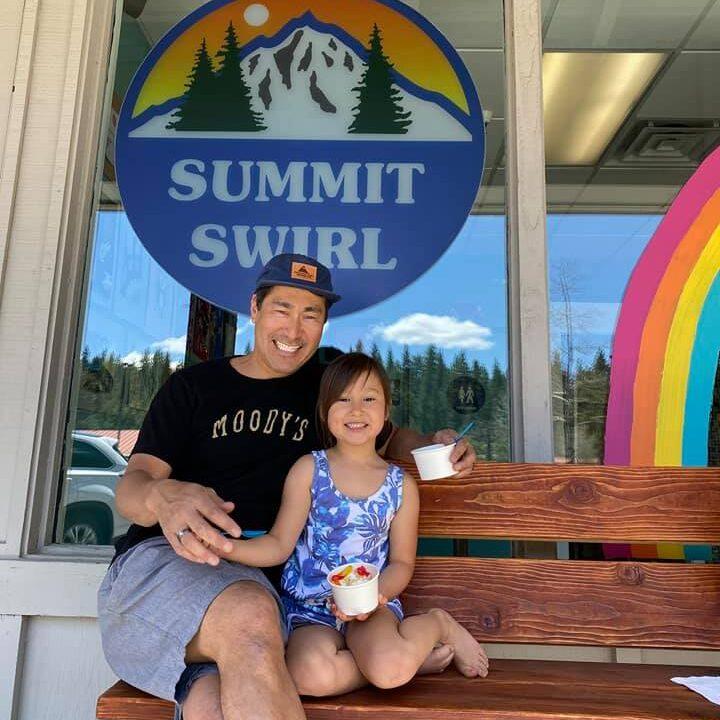 Man and child smiling on bench, holding ice cream outside a shop with rainbow decor.