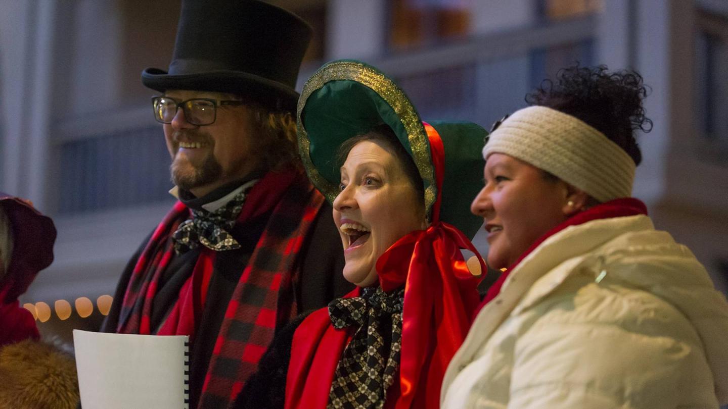 Three carolers in festive attire sing joyfully outside.
