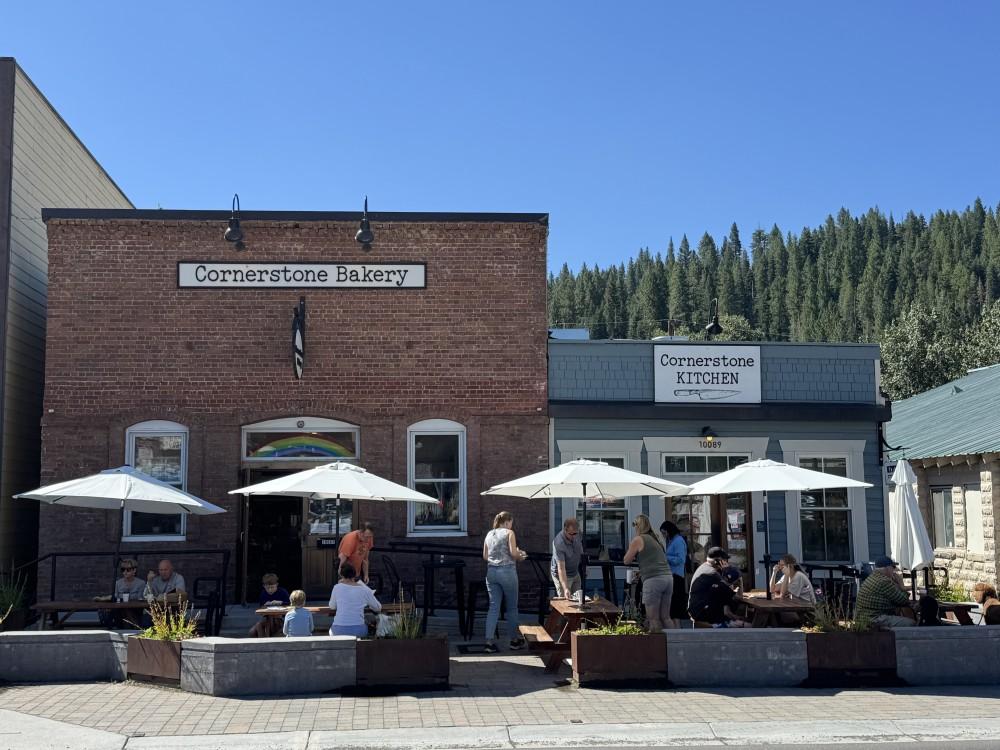 Brick cafe with outdoor seating, umbrellas, and people, against a forested backdrop.