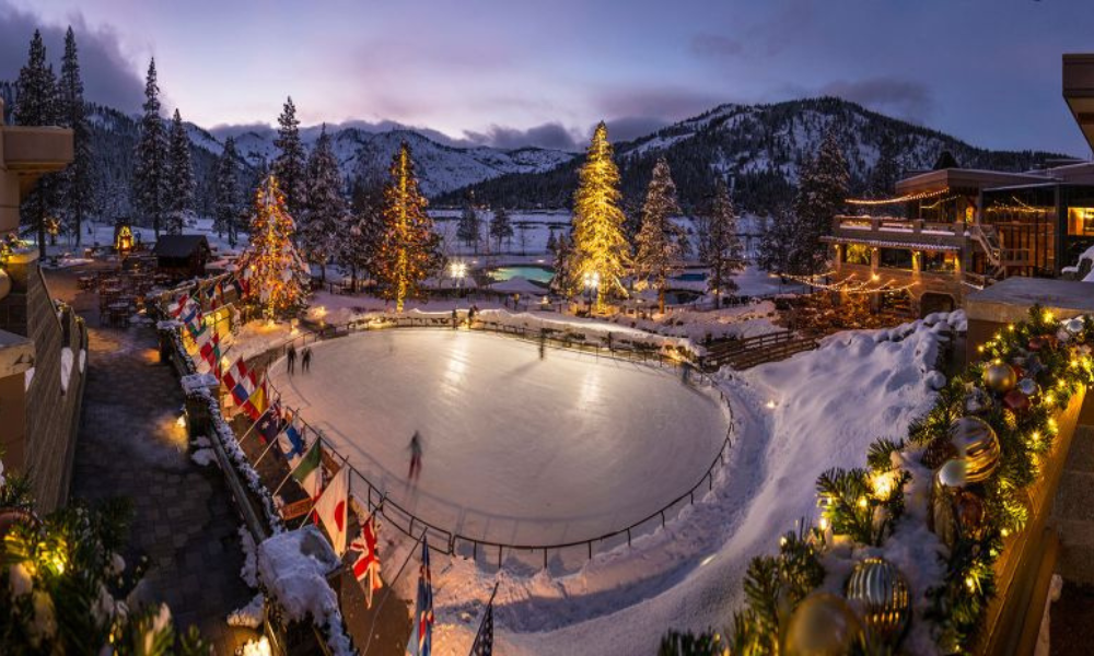 Snowy outdoor ice rink at dusk, surrounded by festive lights and mountains.