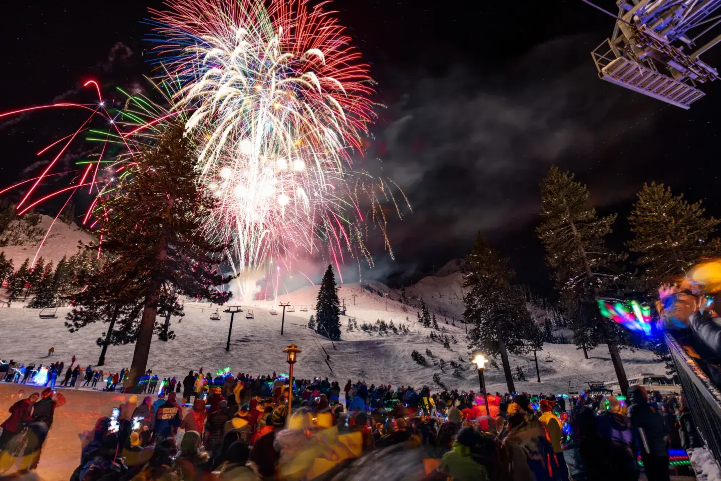 Fireworks light up the night sky above a snowy mountain with a crowd watching.