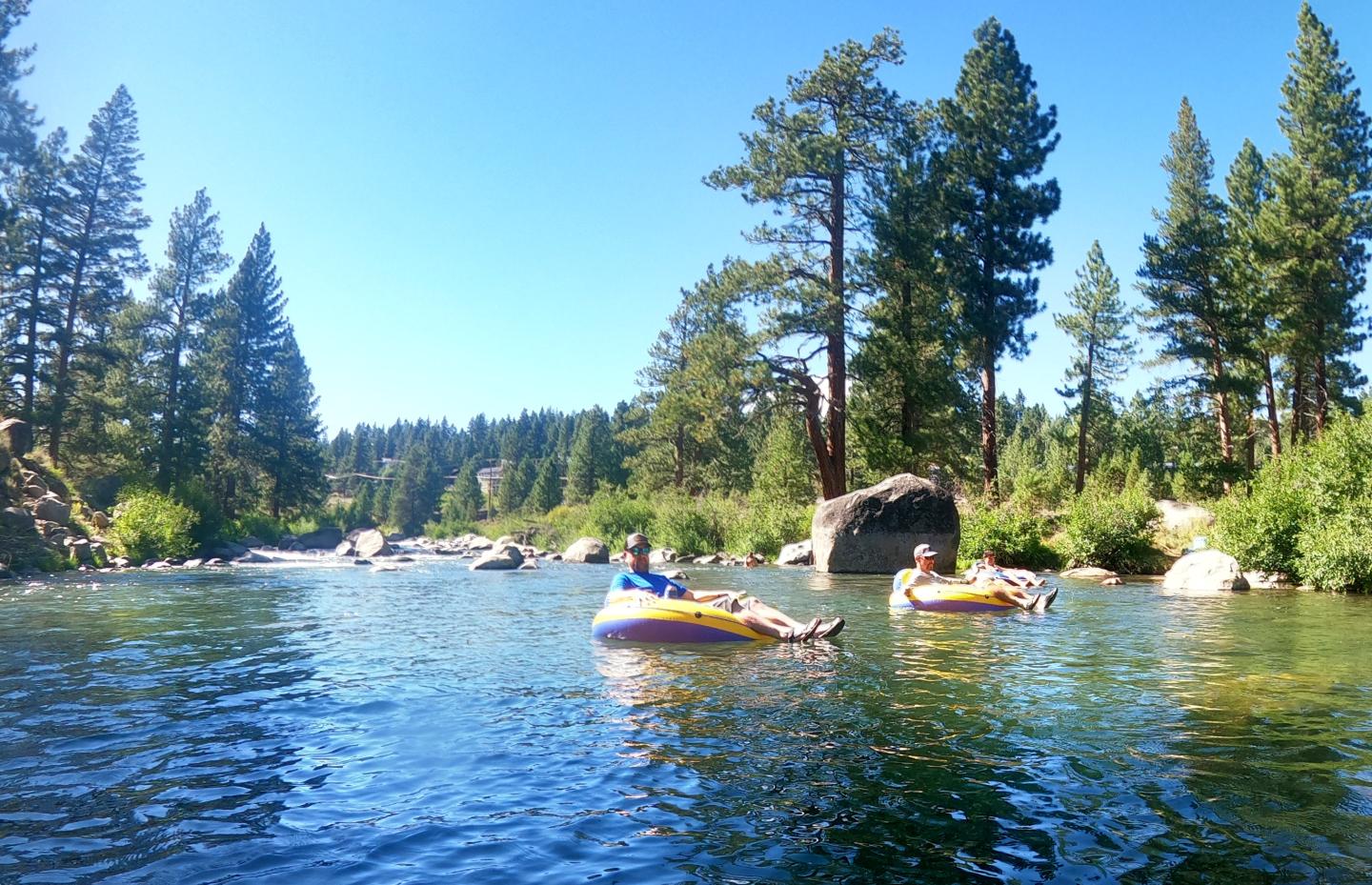 Two people float on a river in inflatable tubes, surrounded by trees on a sunny day.