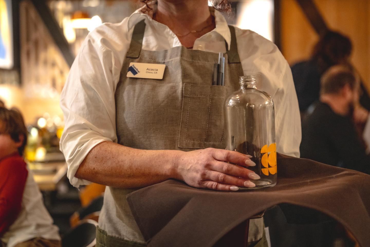 Server carrying a tray with a clear bottle in a restaurant setting.