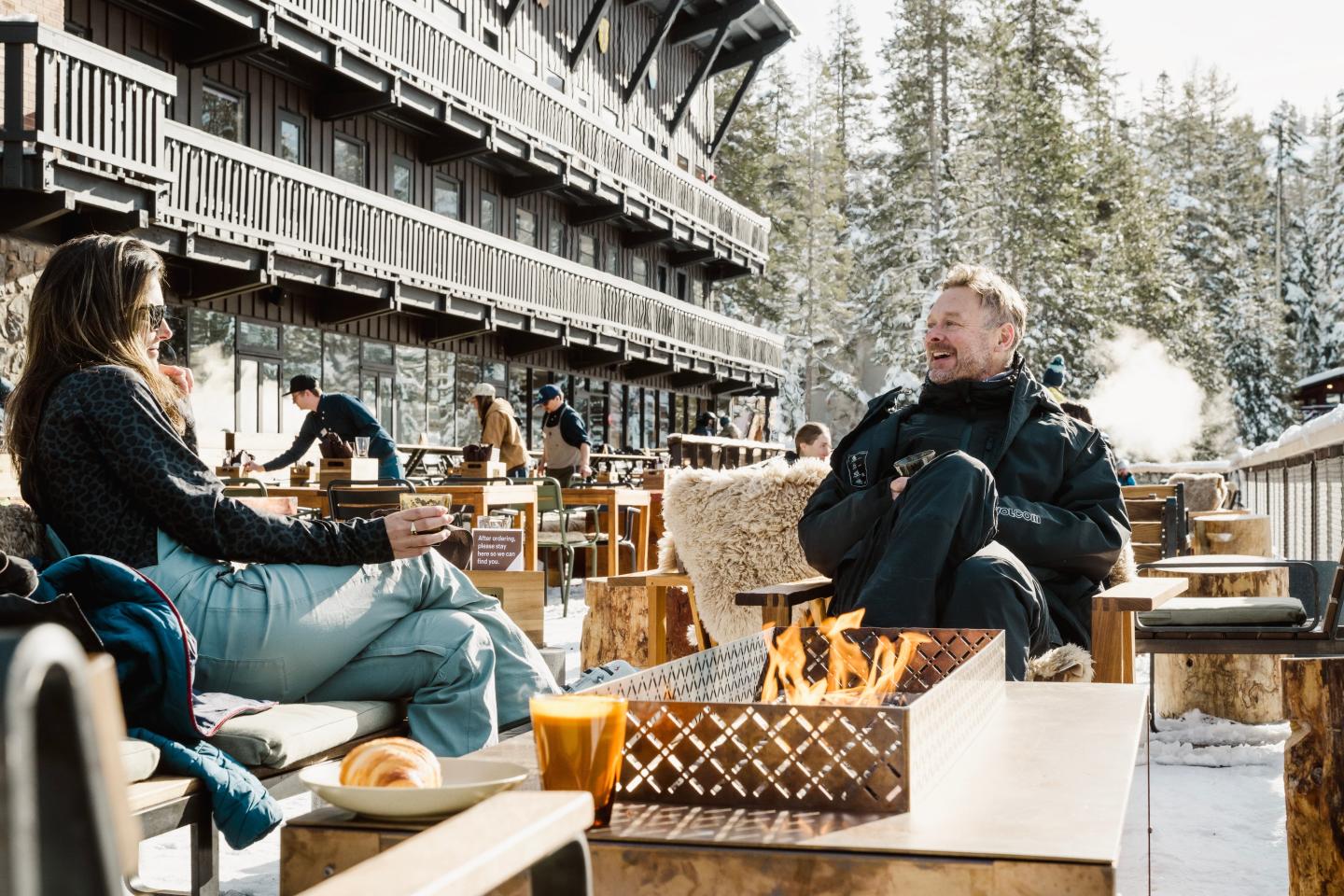 Two people sit by an outdoor fire pit at a snowy mountain lodge.