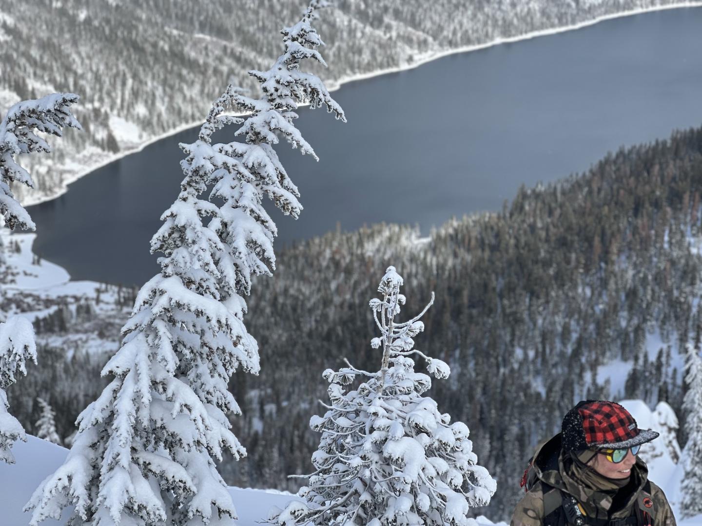 Snow-covered trees on a mountainside overlooking a dark lake, person in winter gear.