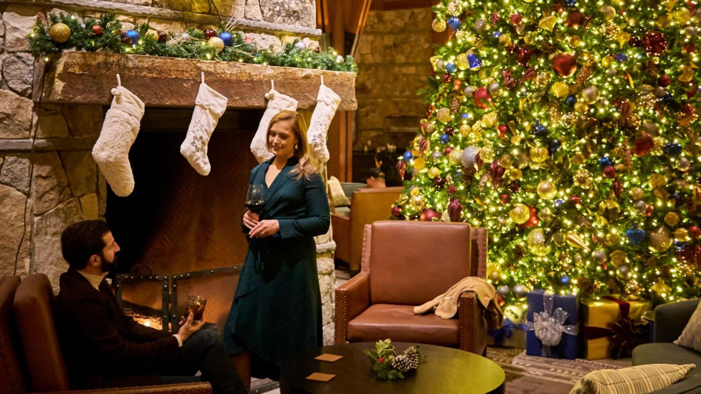 Cozy room with a lit fireplace and decorated Christmas tree, two people talking.