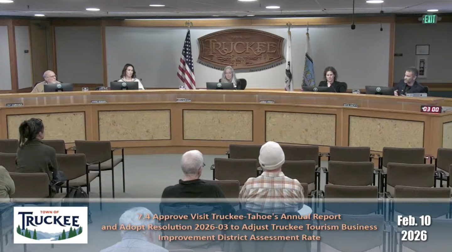 Town council meeting with attendees seated, flag on display, and a wooden emblem on the wall.