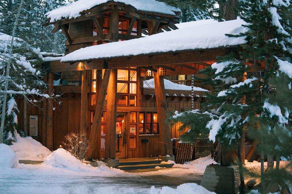 Cozy wooden cabin with snow-covered roof, warmly lit at dusk.