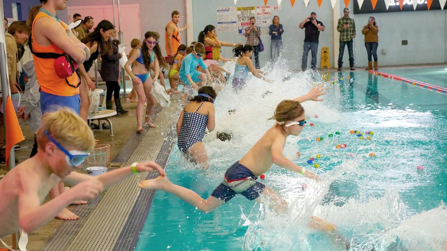 Kids jumping into an indoor pool during a game, splashing water around.