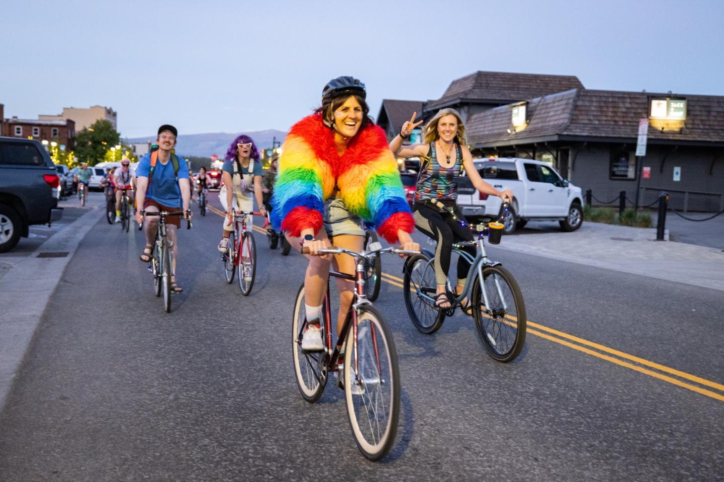 Cyclists in colorful outfits ride bikes on a street, led by a person in a rainbow jacket.