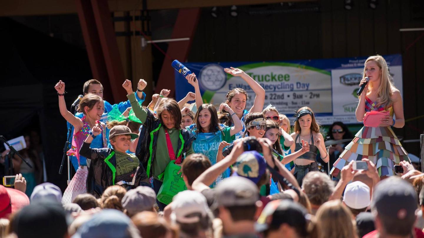 Children cheer on stage during an outdoor event, with a crowd watching.