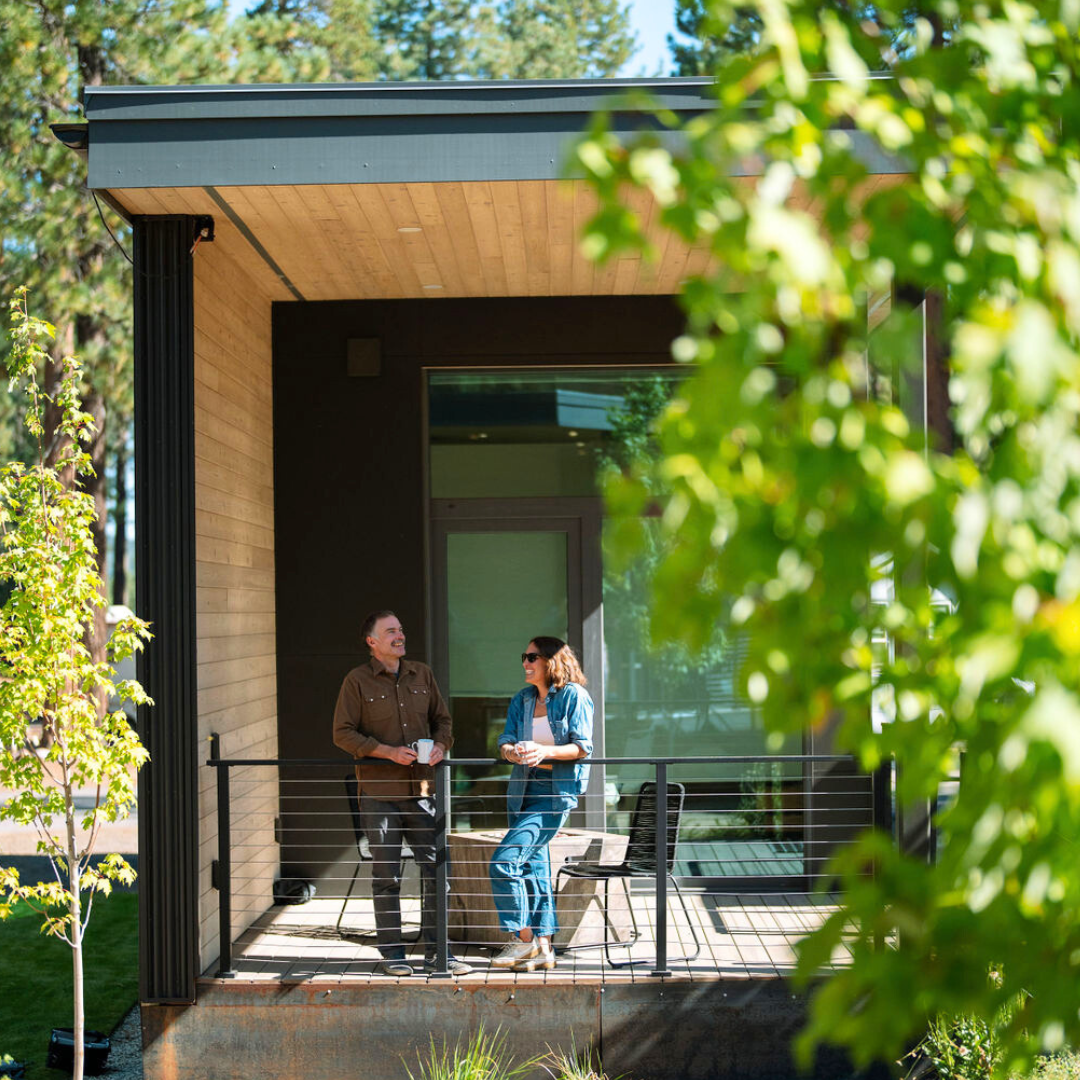 Two people talking on a modern porch, surrounded by green trees.