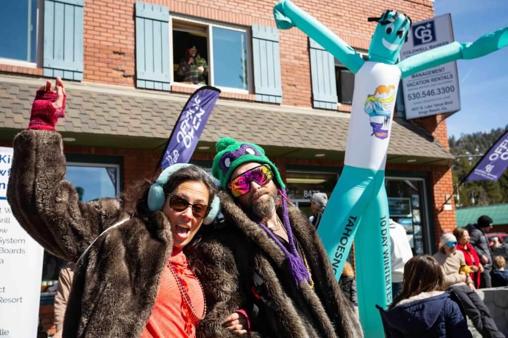 Friends in colorful costumes, joyful at a street festival, inflatable dancer behind.
