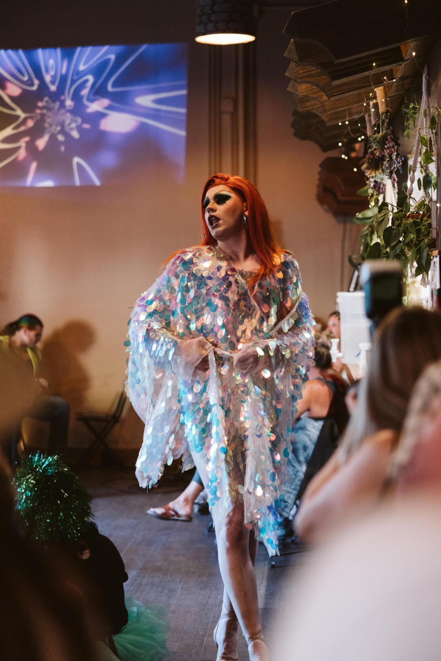 Drag performer in shimmering outfit walks in a lively bar setting.