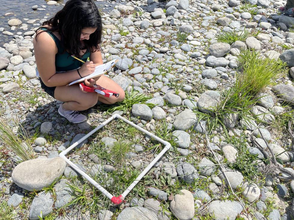 Young woman writing notes by a rock-strewn riverside with a small white frame on the ground.