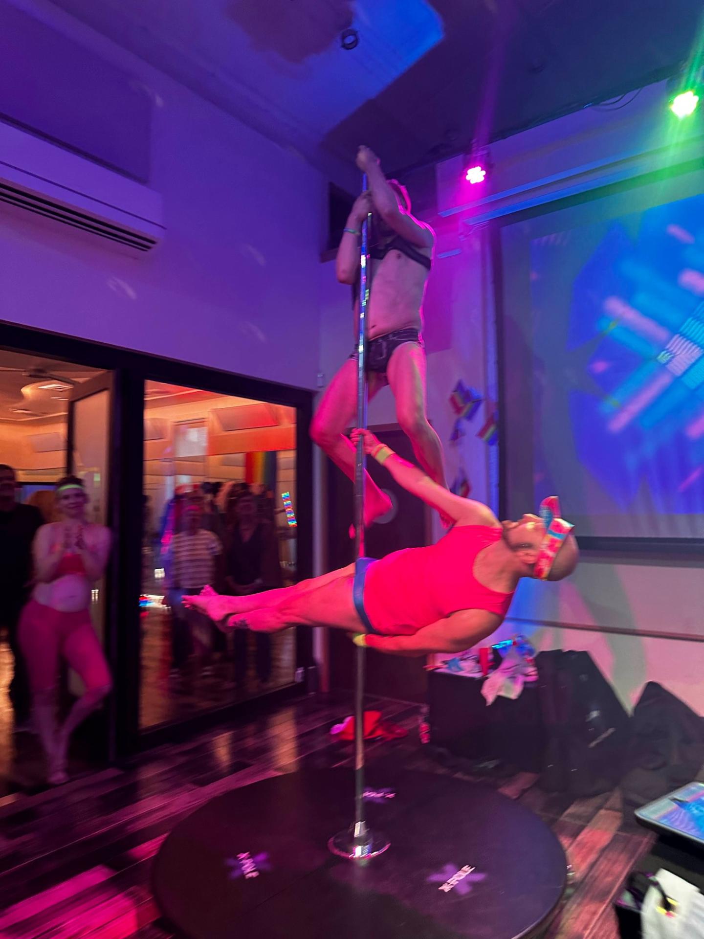 Two performers on a pole under colorful stage lights in a lively venue.