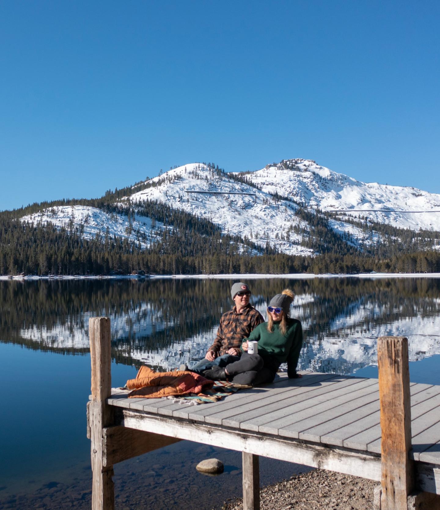 Two people sit on a dock by a lake, snowy mountains in the background.
