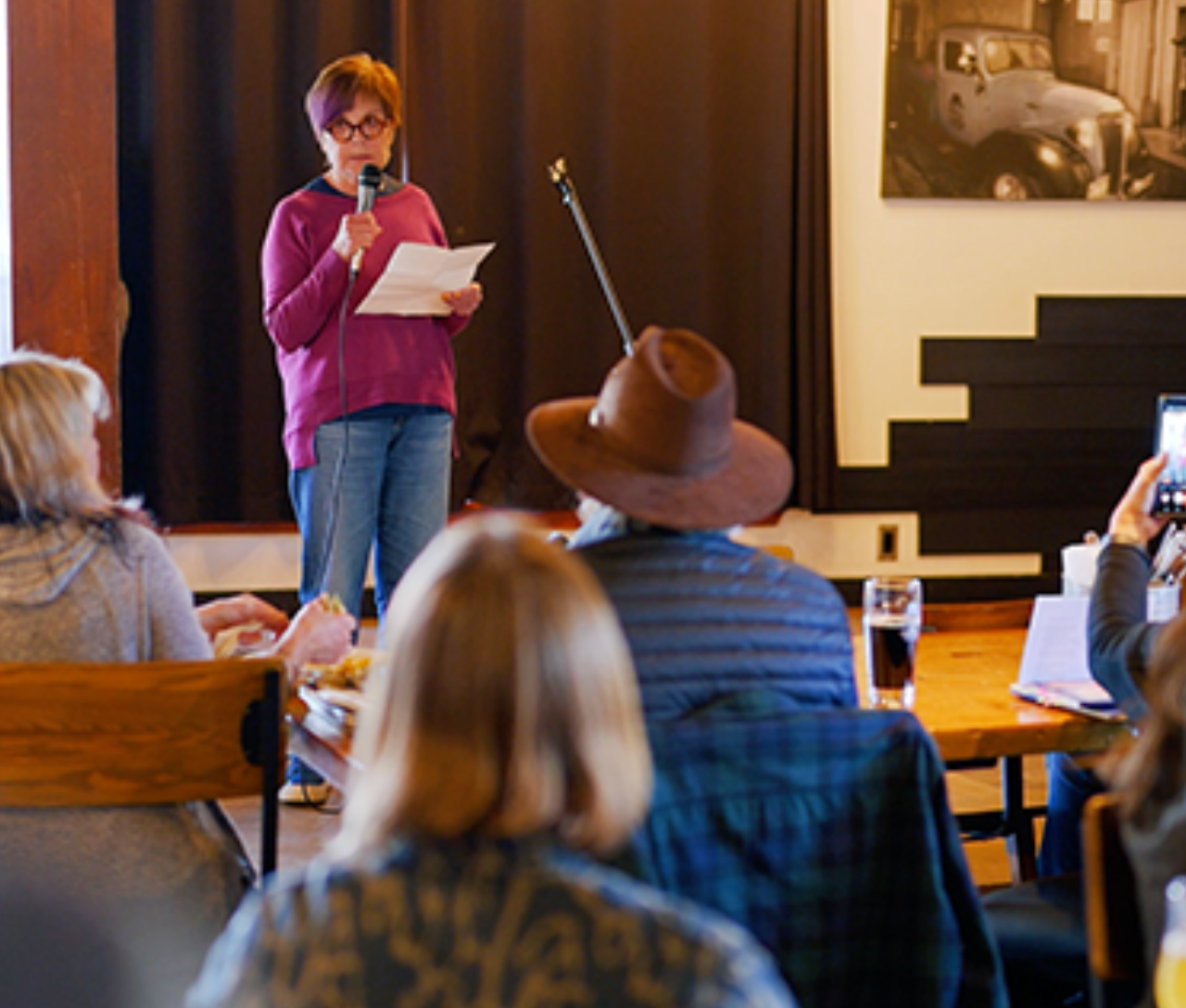 Person with red hair reading on stage, audience seated at tables, cozy setting.