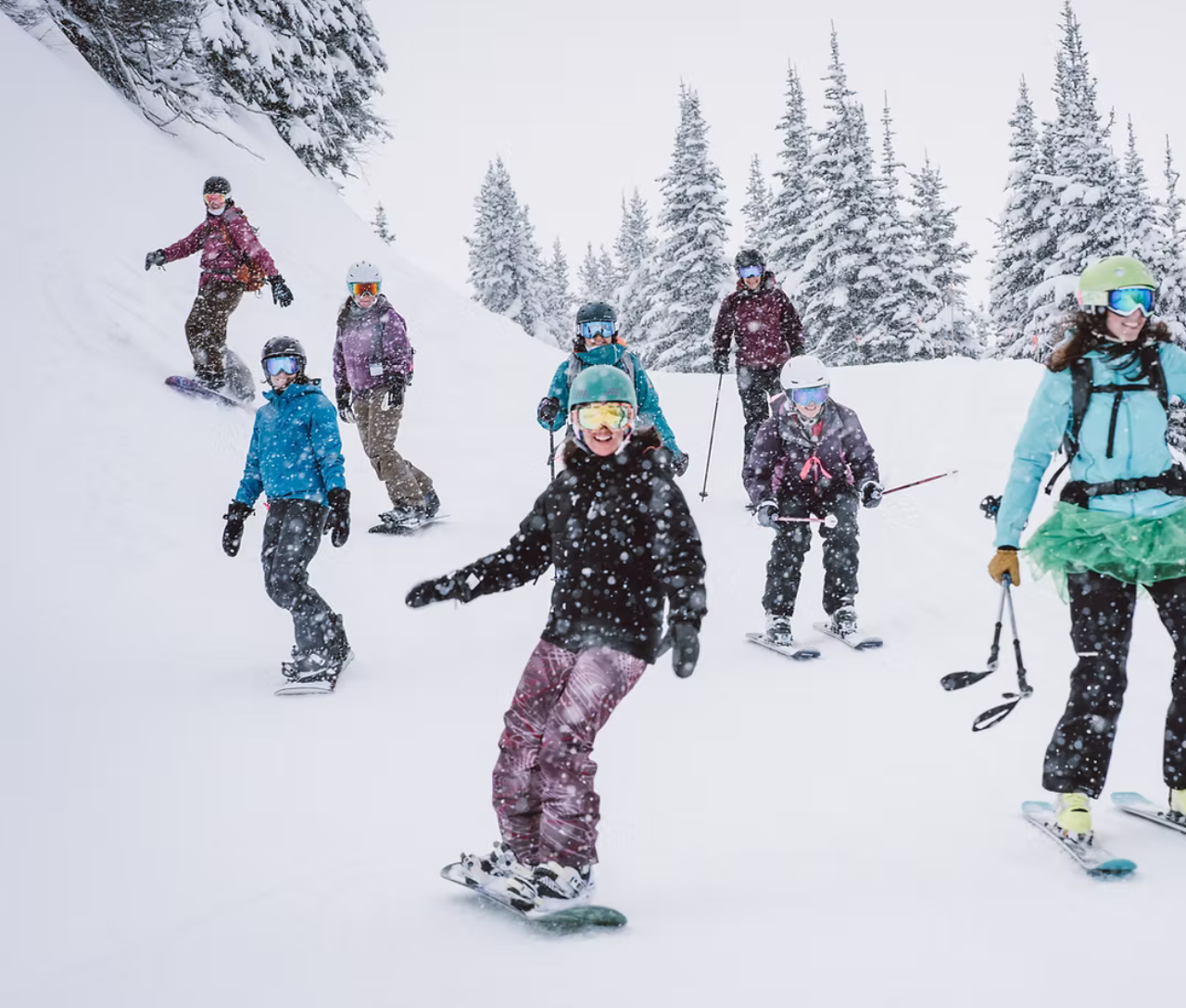 Snowboarders and skiers descending snowy slope, surrounded by pine trees.