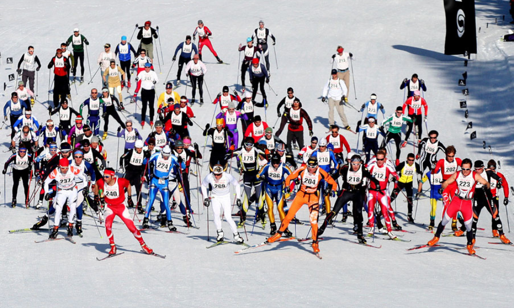 Skiers in a large group race on a snowy trail under clear skies.