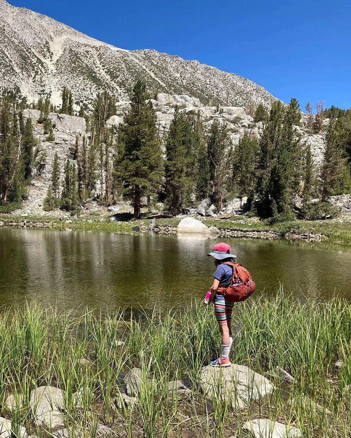 Child with backpack by a mountain lake under clear blue sky.
