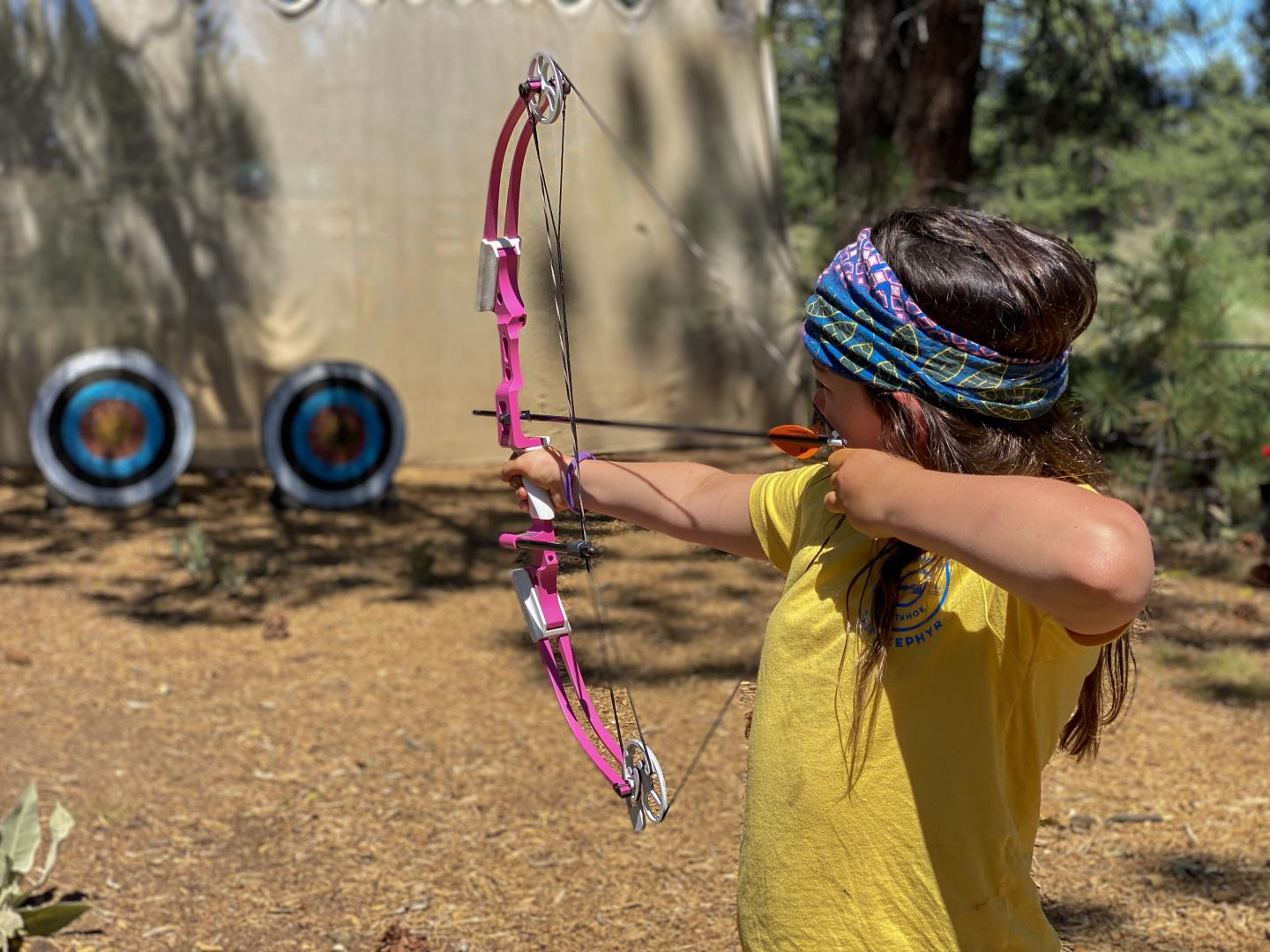 Child aiming pink bow and arrow at targets in an outdoor setting.