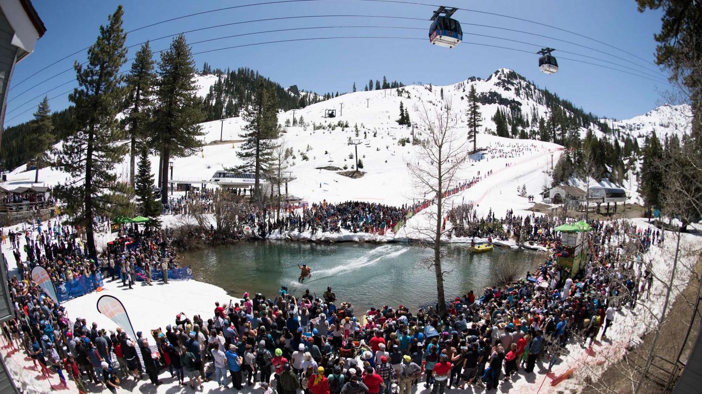 Crowded snowy landscape with pond and skiers, mountains in background.