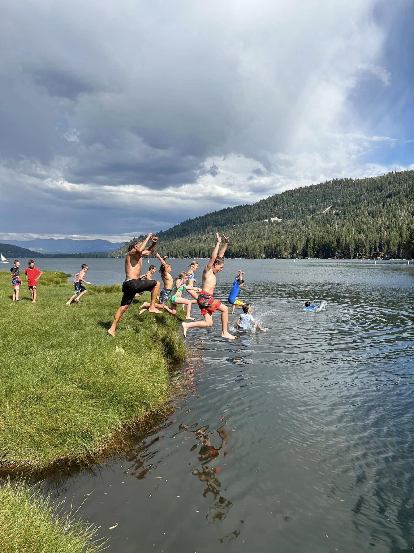 Kids jump into a lake from a grassy bank under a cloudy sky.
