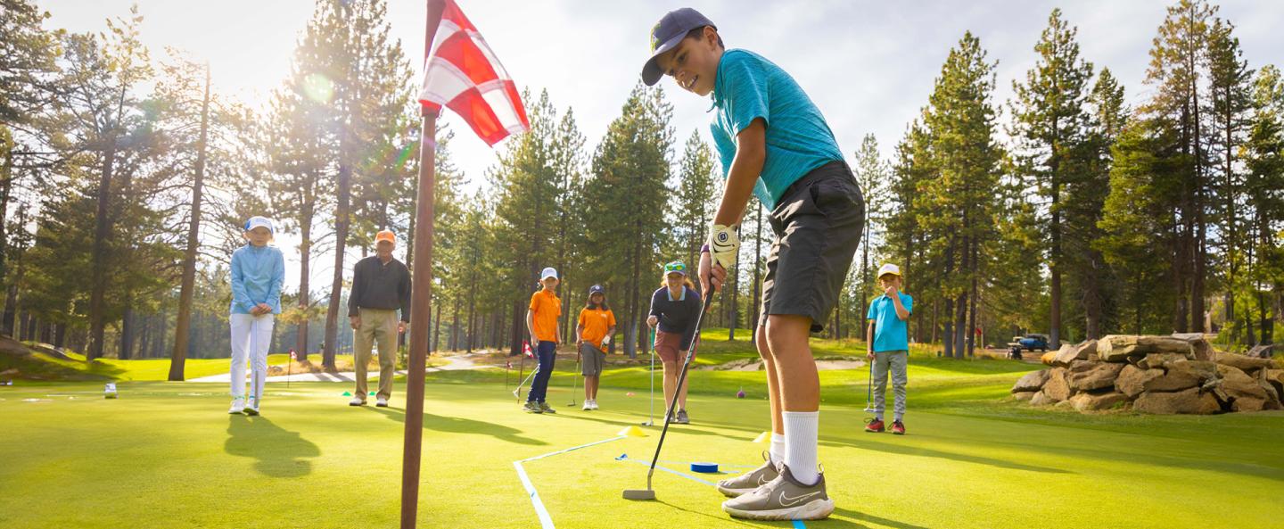 Young boy putting on a sunny golf course, people watching in the background.