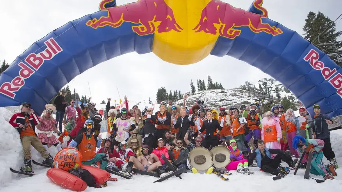 Group of people in colorful costumes under a Red Bull arch on snowy terrain.