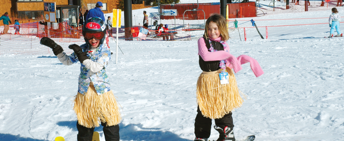 Children in ski gear and hula skirts having fun on a snowy slope.
